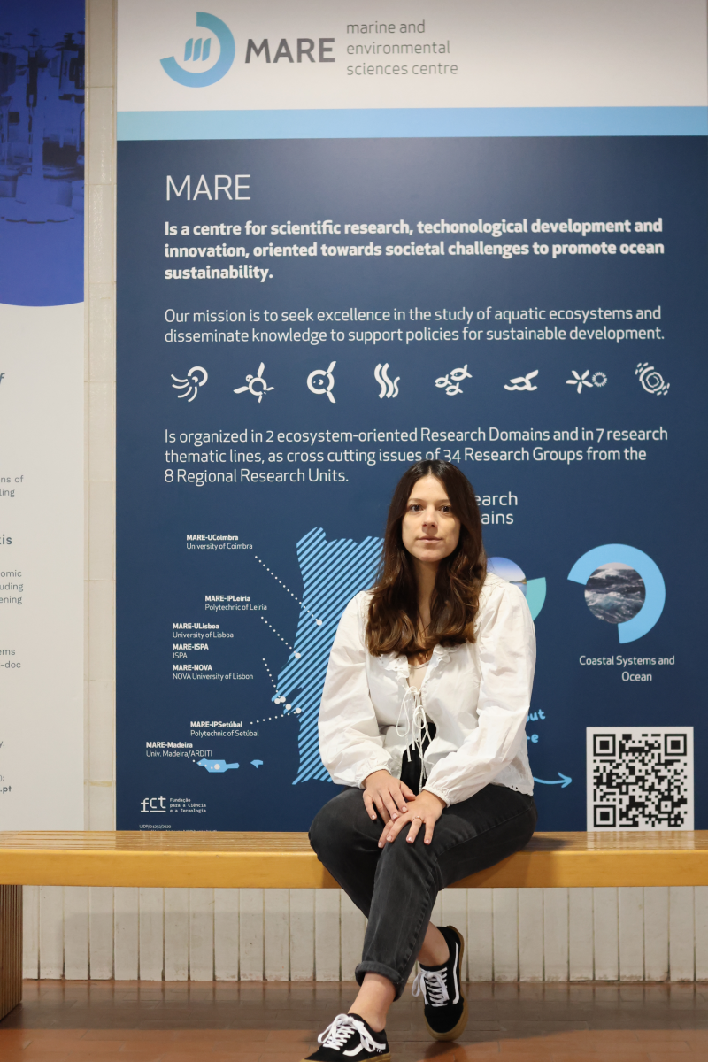 Foto de Ana Sofia Lopes no campus de CIÊNCIAS em frente a poster do MARE.